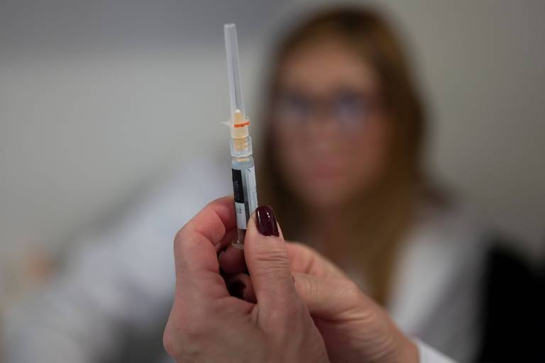 Seringa é segurada por mulher; ao fundo, outra aparece com o rosto desfocado