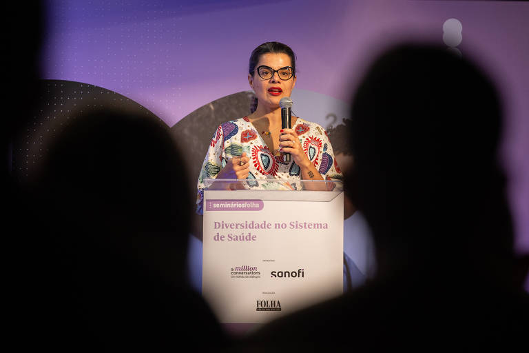 Neila é uma mulher branca de cabelos castanhos, ela está de pé atrás de um púlpito com a inscrição Diversidade no Sistema de Súde