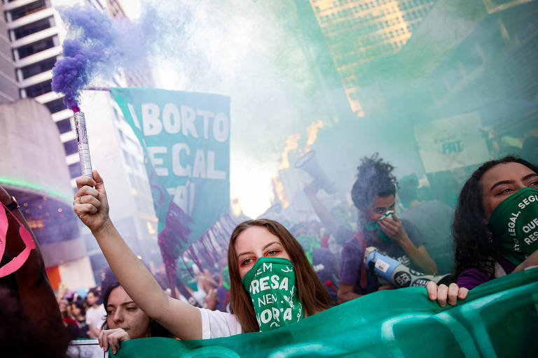 Manifestante com lenço verde cobrindo o rosto segura um sinalizador que solta fumaça roxa