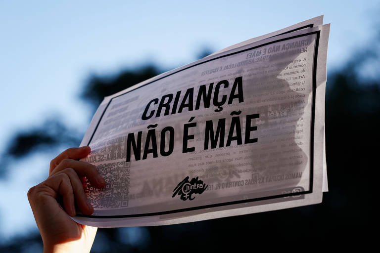 A imagem mostra uma mão segurando um cartaz com a mensagem 'CRIANÇA NÃO É MÃE'. O fundo está desfocado, destacando o cartaz. A mensagem está escrita em letras maiúsculas e em negrito.