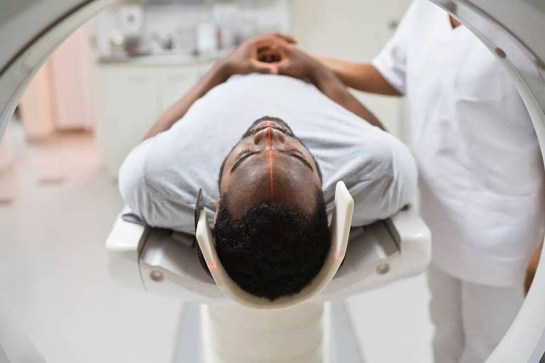 Foto mostra um omem negro entrando em um aparelho branco de tomografia.