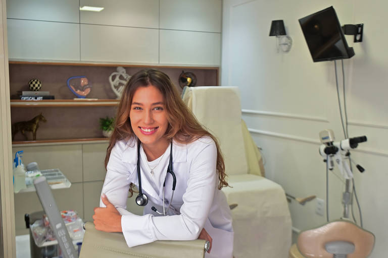 A imagem mostra uma estudante de medicina em um consultório. Ela tem pele branca e cabelos lisos e claro. Ela está usando um jaleco branco, com um estetoscópio ao redor do pescoço. O consultório é bem iluminado, com prateleiras contendo itens decorativos e livros ao fundo. Há também equipamentos médicos visíveis, incluindo uma cadeira de exame médico e um foco de luz.