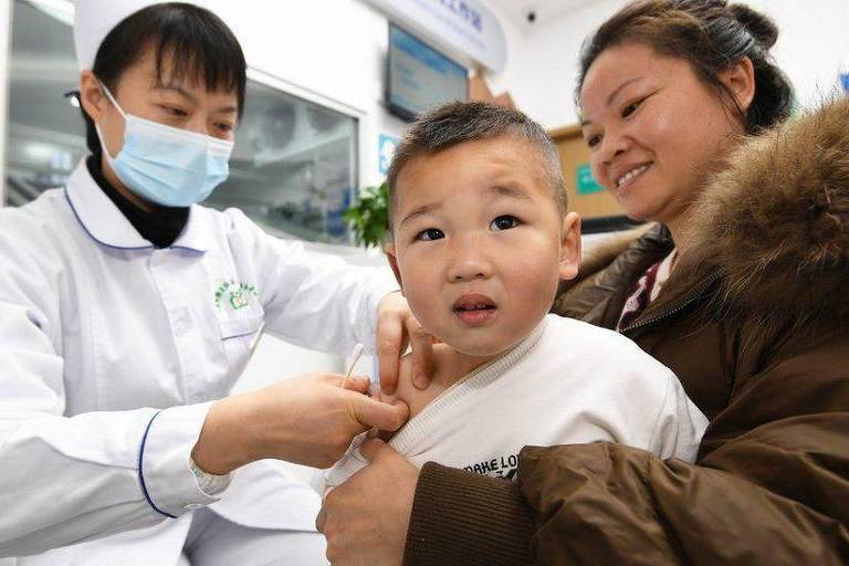 A imagem mostra uma criança pequena, com expressão de preocupação, sendo atendida por uma profissional de saúde que usa um jaleco branco e máscara. A criança está sendo segurada por uma mulher, que parece ser sua mãe, e ambos estão em um ambiente de consultório médico. A mulher está sorrindo para a criança, tentando confortá-la.