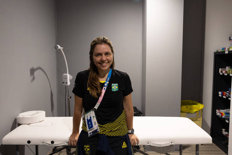 Uma mulher está sorrindo em um ambiente de atendimento, ao lado de uma mesa de tratamento. Ela usa uma camiseta preta com detalhes em amarelo e um crachá pendurado no pescoço. Ao fundo, há uma luz de tratamento e prateleiras com materiais diversos.