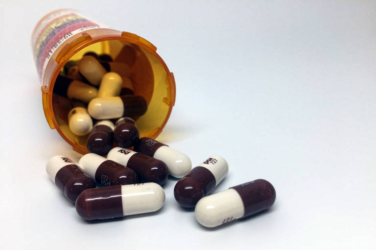 A imagem mostra um frasco de medicamentos derrubado, com várias cápsulas espalhadas ao redor. As cápsulas são predominantemente marrons e brancas, e algumas estão dentro do frasco, que é de cor laranja com uma tampa amarela.
