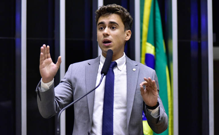 Um jovem homem em um terno cinza e gravata azul está falando em um microfone. Ele gesticula com as mãos enquanto se dirige a uma audiência. Ao fundo, há bandeiras do Brasil visíveis.