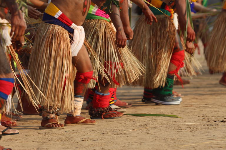 A imagem mostra um grupo de pessoas participando de uma dança tradicional. Os dançarinos estão usando saias feitas de palha e adornos coloridos nas pernas, com faixas vermelhas, azuis e verdes. Os pés estão descalços ou com sandálias, e alguns têm acessórios como pulseiras e fitas. O chão é de terra batida, e a cena captura o movimento dos dançarinos em um ambiente festivo.