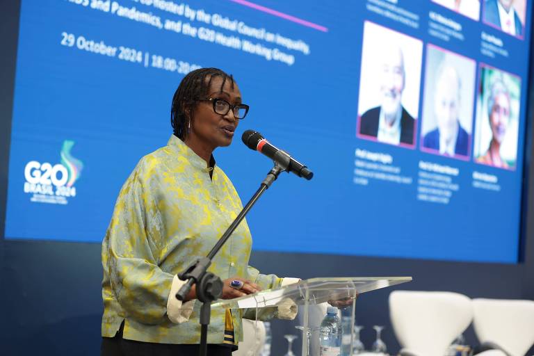 Uma mulher em pé em um palco, falando ao microfone. Ela usa uma blusa verde e óculos. Ao fundo, há um painel com várias fotos e textos relacionados a um evento, datado de 29 de outubro de 2021. O ambiente parece ser uma conferência ou seminário.