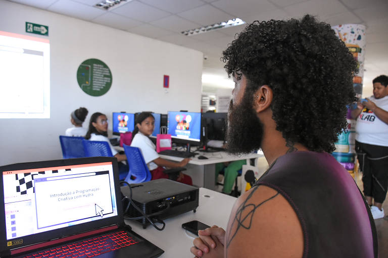 A imagem mostra um ambiente de aula com várias pessoas em computadores. Um homem com cabelo crespo e barba está sentado em frente a um laptop, onde é visível a tela com o texto 'Introdução à Programação Criativa com Hydra'. Ao fundo, há várias crianças sentadas em mesas, algumas olhando para a câmera. Um projetor está exibindo a apresentação na parede. O ambiente parece ser uma sala de aula moderna.