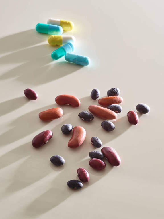 A imagem mostra várias pílulas coloridas em cima de uma superfície clara, com algumas pílulas azuis, amarelas e brancas agrupadas no canto superior. Abaixo, há uma variedade de grãos, incluindo feijões de diferentes cores, como vermelho, marrom e preto, espalhados pela superfície. As sombras dos objetos são visíveis, criando um efeito visual interessante.