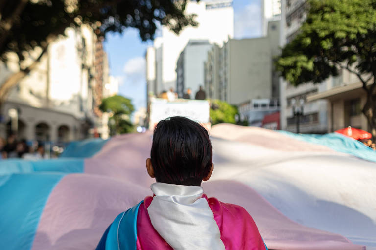 A imagem mostra uma pessoa vista de costas, usando uma capa com as cores da bandeira trans (azul, rosa e branco), em frente a uma grande bandeira trans que se estende pela rua. Ao fundo, há prédios e árvores, indicando um ambiente urbano durante um evento de celebração e protesto.