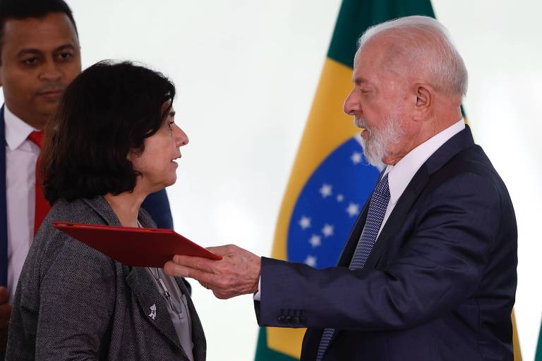 Imagem mostra conversa da ministra Nísia Trindade com o Presidente Lula. Ambos estão de frente um para o outro. Nísia é uma mulher de pele clara e cabelos escuros. Ela vesta um caso cinza. Lula é um homem idoso, grisalho, que veste um terno escuro, com camisa branca. Ele entrega uma pasta vermelha para uma terceira pessoa