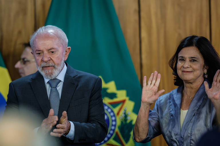 A imagem mostra duas pessoas em um evento oficial. À esquerda, um homem de cabelo grisalho e barba, vestindo um terno escuro e gravata, aplaude. À direita, uma mulher com cabelo escuro e solto, usando uma blusa clara e um casaco azul, acena com as duas mãos. Ao fundo, há bandeiras do Brasil e uma parede de madeira.