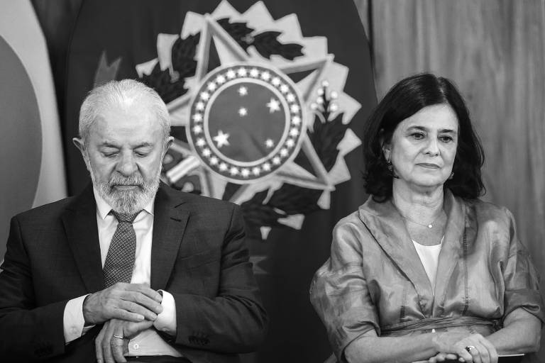 A imagem mostra duas pessoas sentadas em um evento oficial. À esquerda, um homem com cabelo grisalho e barba, vestindo um terno escuro e gravata, está com a cabeça baixa e as mãos cruzadas. À direita, uma mulher com cabelo escuro e liso, vestindo uma blusa cinza, está olhando para frente com uma expressão séria. Ao fundo, há bandeiras do Brasil e um grande símbolo nacional.