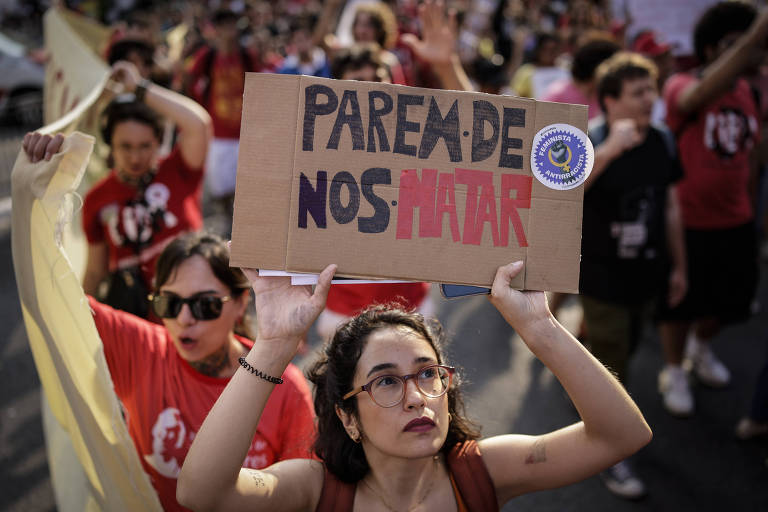 A imagem mostra uma manifestação com várias pessoas. Em primeiro plano, uma mulher com óculos e cabelo solto segura um cartaz que diz 'PAREM DE NOS MATAR'. Ao fundo, outras pessoas também estão participando da manifestação, algumas vestindo camisetas vermelhas.