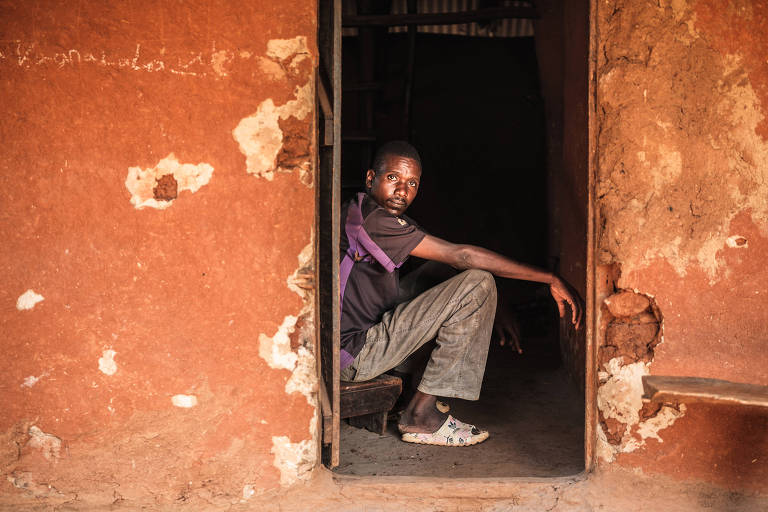 Um jovem está sentado em uma cadeira na entrada de uma casa com paredes de barro. Ele usa uma camiseta preta e calças cinzas, e está com uma perna cruzada sobre a outra. O ambiente é simples, com uma escada visível ao fundo e a luz natural iluminando a cena.