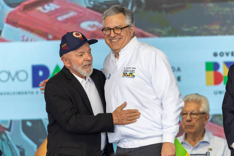 A imagem mostra Lula e Padilha em evento de entrega de ambulâncias do Samu. Lula está está usando um boné com o símbolo do Samu. Padilha, à direita, veste uma camisa branca com logotipos do governo e está sorrindo. Eles estão se cumprimentando, enquanto ao fundo há uma tela com cores e logotipos do governo. Outros homens estão visíveis ao fundo, alguns sentados.