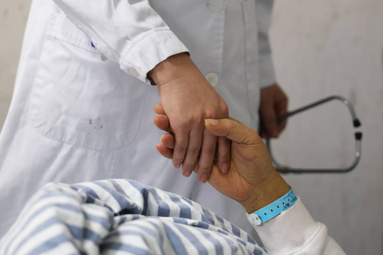 A imagem mostra uma mão de um profissional de saúde, vestindo um jaleco branco, segurando a mão de um paciente que está deitado em uma cama. O paciente usa uma pulseira de identificação azul e está coberto com um lençol listrado em azul e branco. Ao fundo, é possível ver um estetoscópio.