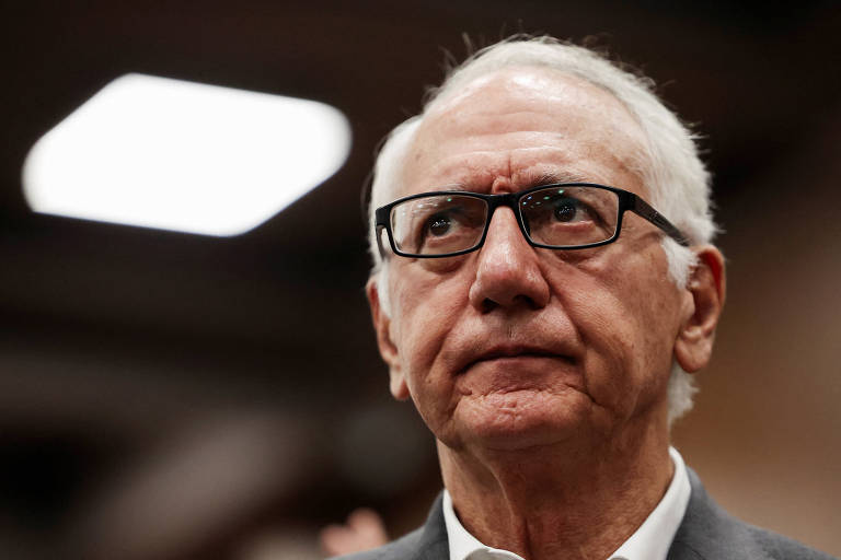 Um homem idoso com cabelo grisalho e óculos, olhando para cima com uma expressão séria. Ele está vestido com uma camisa clara e um paletó escuro. O fundo é desfocado, mas há uma luz quadrada visível no canto superior esquerdo da imagem.