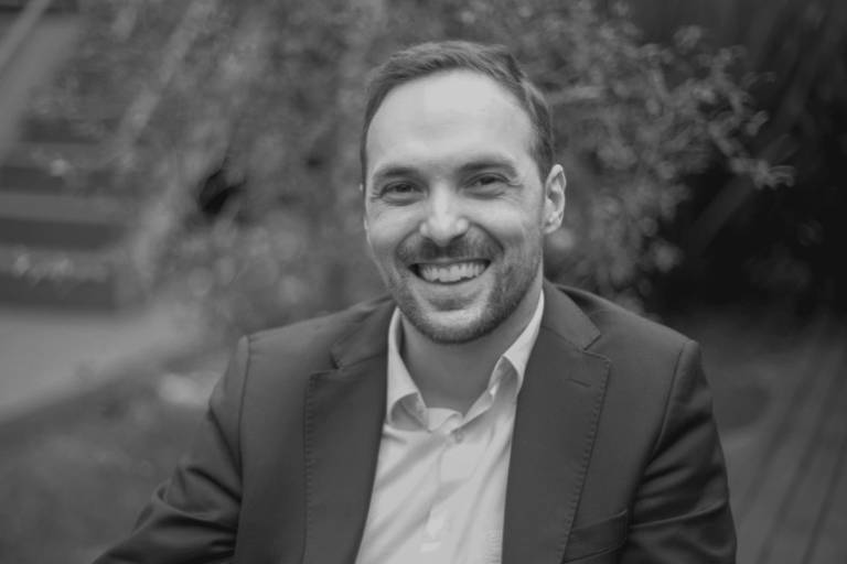 A imagem mostra um homem sorridente, vestido com um terno escuro e uma camisa clara. Ele está sentado em um ambiente externo, com um fundo desfocado que sugere um espaço ao ar livre. O homem tem cabelo curto e uma barba bem aparada. A foto é em preto e branco, destacando a expressão amigável do homem.