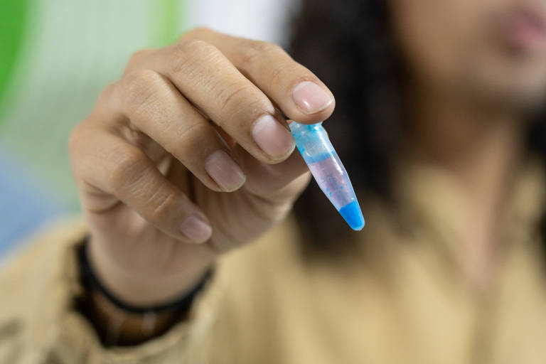 A imagem mostra uma mão segurando um tubo de ensaio pequeno, que contém um líquido azul na parte inferior e um líquido transparente na parte superior. O fundo é desfocado, mas parece ter cores verdes e azuis.