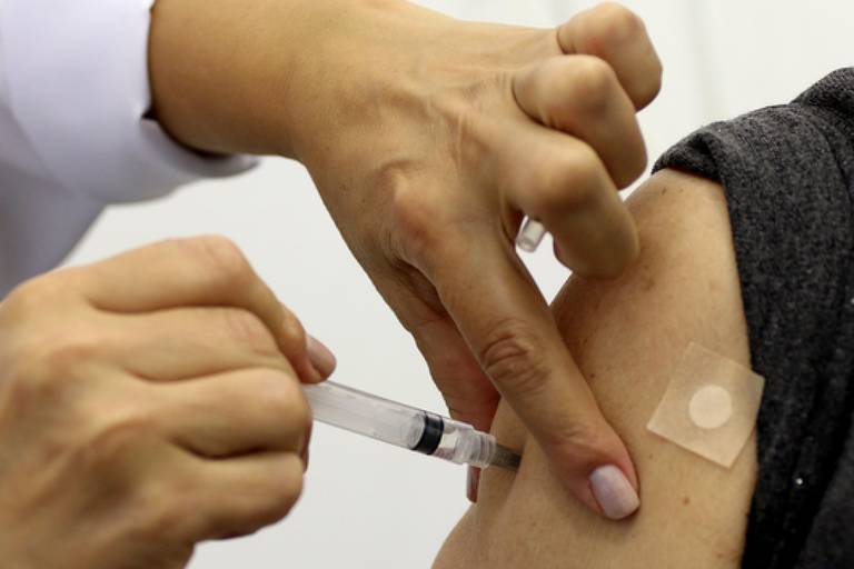 A imagem mostra uma mão segurando uma seringa enquanto outra mão está aplicando a vacina em um braço exposto. O braço tem um adesivo sobre o local da injeção, e a pessoa que aplica a vacina está usando um jaleco branco.