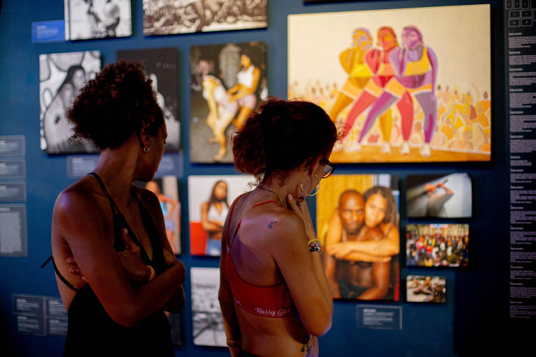 Duas mulheres estão observando obras de arte em uma exposição. Elas estão de costas, uma com cabelo cacheado e a outra com cabelo liso. As paredes ao fundo estão cobertas com várias fotografias e ilustrações, incluindo uma pintura colorida de quatro figuras femininas em trajes esportivos. A iluminação é suave, criando um ambiente contemplativo.