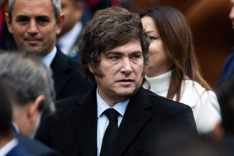 A imagem mostra um homem com cabelo castanho e ondulado, vestindo um paletó preto e uma gravata preta. Ele parece estar em um evento público, cercado por outras pessoas. Ao fundo, é possível ver rostos de outras pessoas, algumas com expressões neutras. O ambiente parece ser ao ar livre, com uma multidão ao redor.