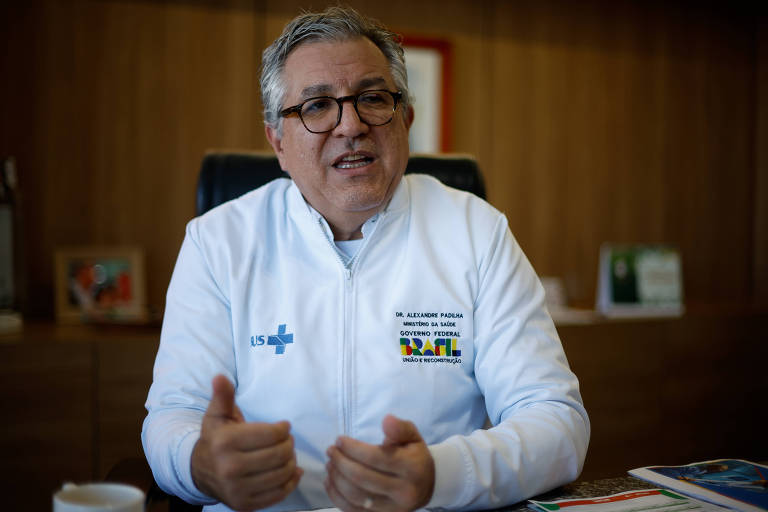 Um homem com cabelo grisalho e óculos está sentado em uma cadeira, usando um jaleco branco com o logotipo de uma instituição de saúde. Ele está gesticulando com as mãos enquanto fala. Ao fundo, há uma mesa de escritório com alguns objetos e documentos, além de uma parede de madeira. O ambiente parece ser um escritório.