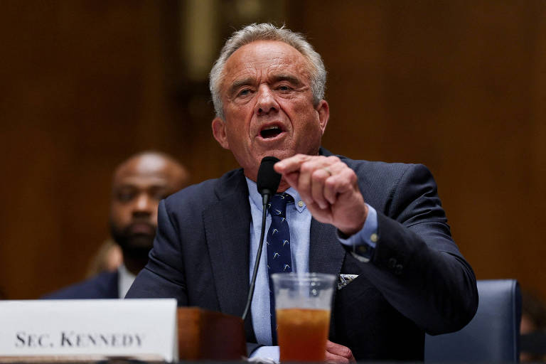Um homem de cabelo grisalho e com um terno escuro está falando em um microfone durante uma audiência no Senado. Ele parece estar expressando uma opinião forte, com a mão levantada. Ao fundo, há um outro homem sentado, observando. Na mesa à sua frente, há um copo com uma bebida e um nome em uma placa que diz 'SEC. KENNEDY'.