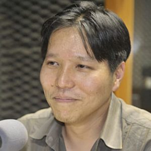 foto de um homem oriental, cabelos pretos curtos e olhos pretos. Ele está falando ao microfone e, ao fundo, existe uma parede com espumas anti-ruído.