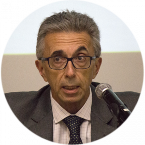 Homem branco, idoso, usando óculos e vestindo paletó escuro, camisa clara e gravata preta com bolinhas brancas