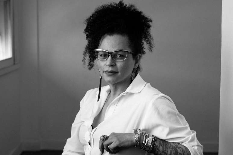 Imagem em preto e branco mostra Neon Cunha, uma mulher negra, de pele clara, que veste uma camisa branca e está sentada.