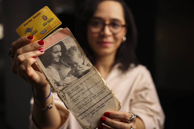 Uma mulher está segurando um documento antigo que contém uma foto em preto e branco de duas pessoas. O documento parece estar desgastado e possui anotações manuscritas. Acima do documento, há um cartão de identificação com informações visíveis. A mulher tem cabelo escuro, usa óculos e está vestida com uma blusa clara. Ela sorri levemente enquanto segura o documento.